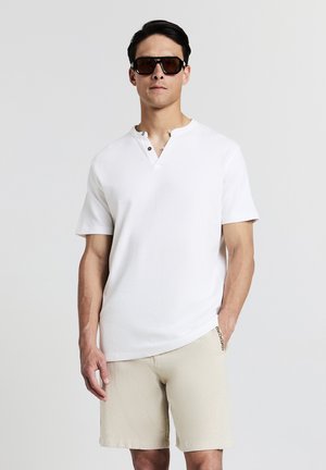 Camicetta henley bianca a maniche corte con due bottoni marroni al colletto, abbinata a pantaloni corti beige texturizzati. Modello con occhiali da sole scuri.