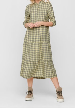 Vrouw draagt een gele geruite midi-jurk met lange mouwen en beige enkellaarsjes, staand met gespreide benen en handen voor zich gesloten.
