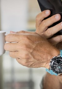 Srebrny zegarek z czarną tarczą i turkusowym silikonowym paskiem, trzymany w dłoni, która chwyta małą białą filiżankę. Tło jest rozmyte.