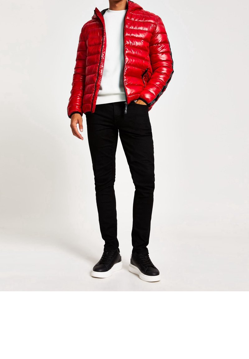 Doudoune rouge à finition brillante, avec des poignets côtelés, des accents noirs sur les côtés, associée à un jean noir et des baskets noires avec des semelles blanches.