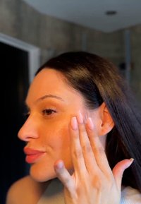 En person med långt mörkt hår applicerar produkt på sin kind. Huden är slät med en naturlig lyster, och naglarna är manikyrerade med en ljus färg.