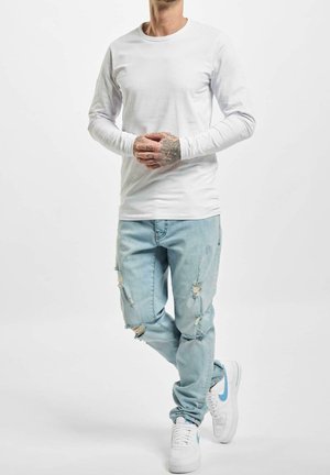 Uomo con camicia bianca a maniche lunghe, jeans azzurri strappati e scarpe da ginnastica bianche con swoosh blu, in piedi con le mani unite.