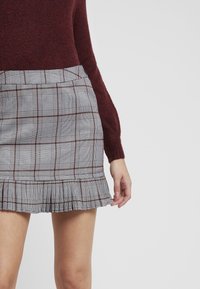 Jupe à carreaux grise, rouge et blanche avec un ourlet plissé. Le matériau semble tissé avec une texture lisse. Accompagnée d'un pull bordeaux.