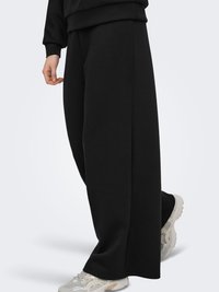 Persoon draagt losse zwarte broek en witte chunky sneakers, staand tegen een effen lichte achtergrond.