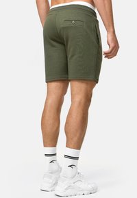 Olivgrüne Shorts mit einer lockeren Passform, elastischem Bund, Gesäßtasche mit Knopf und Seitentaschen. Getragen mit weißen Socken und Schuhen.
