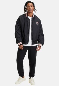Bomber negro con puños de canalé a rayas rojas y blancas, usado sobre una camisa blanca, combinado con joggers negros que presentan un logotipo bordado.