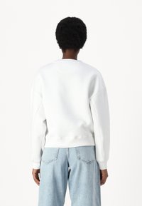 Felpa bianca con scollatura rotonda, spalle scese e polsini a coste. Abbinata a jeans di denim azzurri, mostrata da dietro.