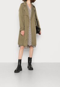 Manteau trench vert olive à coupe cintrée, coupe croisée, texture lisse. Associé à une robe fleurie et des bottines noires.