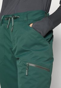 Groene technische broek met een gemêleerd accent, voorzien van een trekkoord in de taille, zijzakken en een ritszak op het linkerbeen.