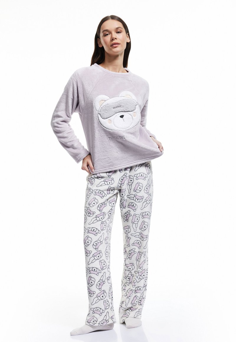 Parte superior de pijama morada y esponjosa con una cara de oso y una máscara de dormir. Pantalones blancos a juego con un estampado repetido de ositos de peluche en colores pastel.