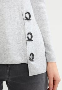 Szary sweter dzianinowy z trzema dużymi srebrnymi akcentami w kształcie pierścienia po boku. Miękka faktura, żebrowany wzór i lekko zaokrąglony dół.