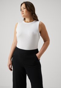 Débardeur blanc à côtes avec un design sans manches, associé à un pantalon noir taille haute avec des poches latérales. Accent avec un collier en chaîne dorée.