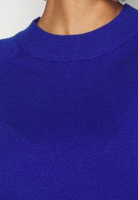 Nära bild av en person som bär en klarblå stickad tröja med rund halsringning mot en enfärgad bakgrund.