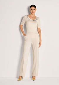 Vrouw staande tegen de muur in een crèmekleurige top met versieringen en een ronde hals, bijpassende hoge taille broek, gouden hakken en lange oorbellen.