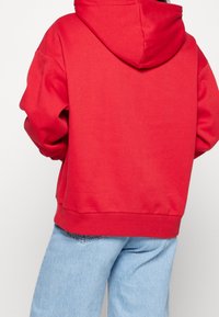 Czerwona bluza z kapturem i sznurkiem, charakteryzująca się luźnym krojem oraz ściągaczami na pasku i rękawach. Materiał wydaje się miękki i teksturowany.