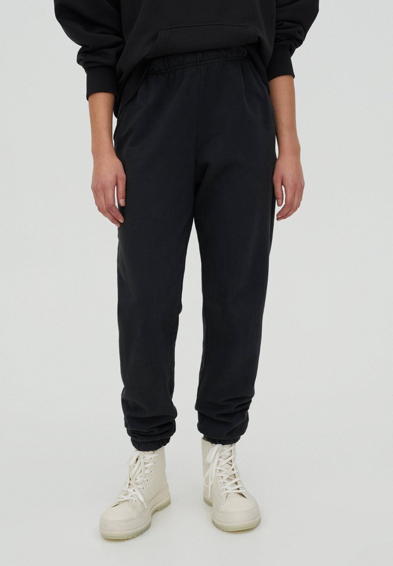 Personne portant un pantalon de jogging noir et des bottines blanches à lacets, debout devant un fond clair uni.