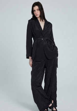 Femme aux cheveux foncés porte un blazer noir texturé ceinturé et un pantalon à jambes larges, debout devant un fond gris clair uni.