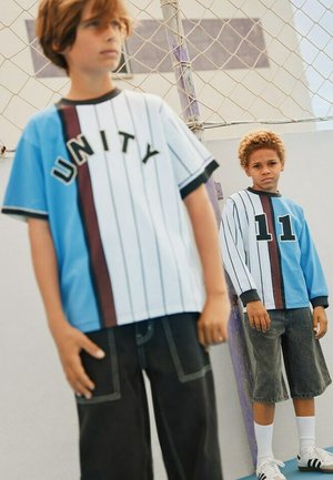 Niebiesko-biała koszula w paski na krótki rękaw z napisem "UNITY", zestawiona z czarnymi szortami. Druga postać ma na sobie niebieską koszulkę z siateczki z napisem "11".