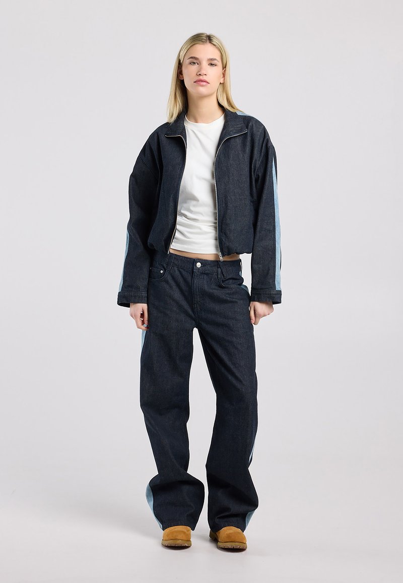 Giovane donna in piedi che indossa una giacca di jeans scura e jeans con strisce laterali chiare, camicia bianca e scarpe di camoscio marrone su sfondo uniforme.