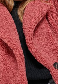 Miękka, teksturowana odzież wierzchnia w głębokim koralowym kolorze, z podwójnym rzędem czarnych guzików, noszona na czarnym prążkowanym swetrze.