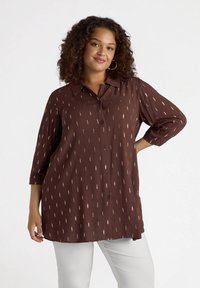 Camicia marrone con bottoni, dotata di colletto, caratterizzata da strisce verticali dorate. Il tessuto appare leggero con una consistenza liscia e maniche a tre quarti.