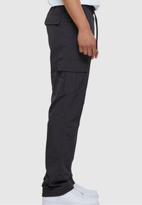 Černé cargo kalhoty z látky s jemným leskem, s postranními kapsami, elastickým pasem a zúženým střihem. Nošeny s bílými teniskami.