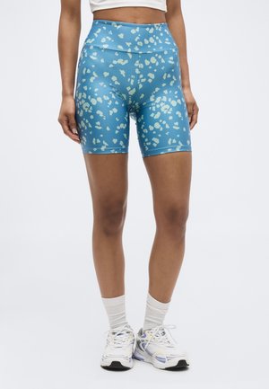 Kvinde iført blå højttaljede cykelshorts med mønster, hvid cropped top, hvide sokker og hvide løbesko på ensfarvet baggrund.