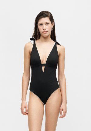 Žena s dlhými tmavými vlasmi má na sebe čierne jednodielne plavky s hlbokým výstrihom do V a bielymi ramienkami zviazanými na mašľu na svetlom jednoliatom pozadí.