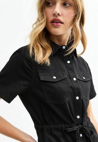 Black short-sleeve shirt dress made of cotton, featuring two chest pockets, button front, and belted waist detail. Smooth texture.