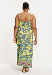 Robe maxi en tissu jaune avec des motifs floraux bleus, dos froncé, fines bretelles et ourlet à motifs. Portée avec des sandales à talons carrés beiges.