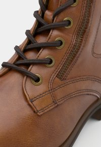 Närbild på brun skinnstövel med mörkbruna snören trådade genom mässingsöglor och sidodetalj med blixtlås.