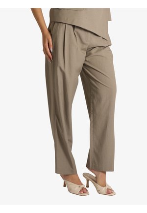 Donna che indossa pantaloni beige plissettati e sandali beige con tacco e cinturini incrociati, in piedi davanti a uno sfondo bianco.