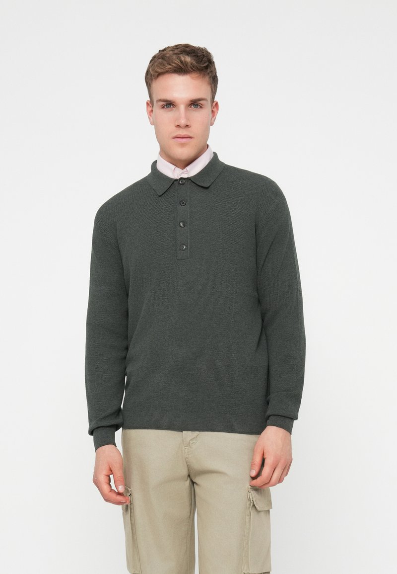 Polo à manches longues de couleur vert foncé avec un tricot texturé, présentant un col, une patte de boutonnage à trois boutons et des poignets côtelés. Porté avec un pantalon cargo beige.
