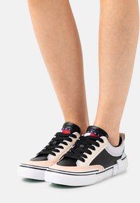 Czarne i beżowe niskie sneakersy wykonane z materiałów skórzanych i tkaninowych, z czarnymi sznurowadłami, białymi podeszwami i kolorowym logo na języku.