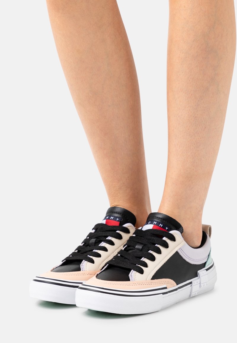 Czarne i beżowe niskie sneakersy wykonane z materiałów skórzanych i tkaninowych, z czarnymi sznurowadłami, białymi podeszwami i kolorowym logo na języku.