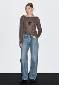 Hnědý pletený svetr se širokým výstřihem, světle modré džíny široké nohavice a černý náhrdelník s přívěskem; model stojí na jednobarevném pozadí.