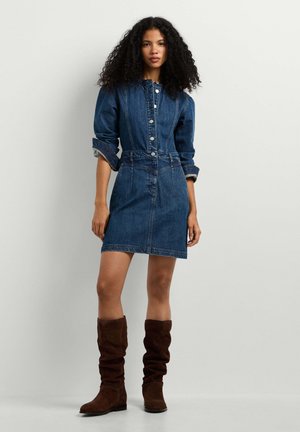 Vrouw met krullend haar, gekleed in een donkerblauwe denim jurk met knopen en bruine slouchy kniehoge laarzen, staand tegen een eenvoudige lichte achtergrond.