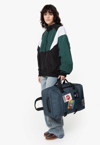 Dark teal travel bag with black straps and multiple patches. The bag has a rectangular shape and a textured fabric. Worn with light blue jeans.