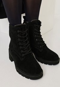 Black suede ankle boots with a lace-up front, a side zipper, textured surface, and block heels. Features stitching details and rubber soles.