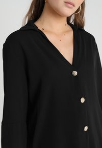 Svart skjorta med knappar, v-ringning och krage, med guldtonade knappar, mjuk struktur och långa ärmar.