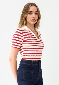 Sweter polo dzianinowy w czerwono-kremowe poziome paski, z krótkimi rękawami i dekoltem w kształcie litery V. W zestawie z dżinsami o wysokim stanie w ciemnym kolorze.