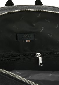 Interno di uno zaino in tessuto nero con una tasca con zip, etichetta in pelle con la scritta "BOSS" e un sottile motivo a logo ripetuto. Dettagli della zip in argento.