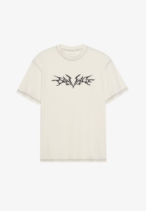 T-shirt in cotone crema con vestibilità rilassata, caratterizzato da testo "SAVAGE" in stile tribale nero al centro. Cuciture a contrasto lungo i bordi.