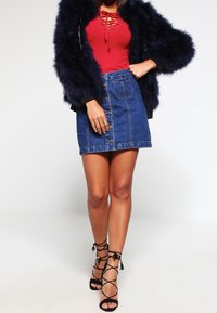 Červený žebrovaný bodysuit s šněrováním vpředu, námořnický umělý kožíšek, džínsová mini sukně s předními knoflíky a černé sandály na vysokém podpatku se zavazováním.