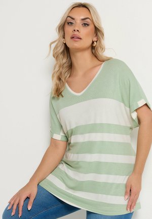Blonde woman wearing gold hoop earrings and a loose mint green and white striped short-sleeve top, posing against a white background.