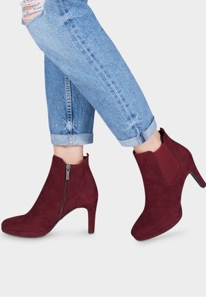 Bourgognefarvede ankelstøvler i ruskind med høj hæl, sidesyning med lynlås og elastisk panel; kombineret med opkørte lyseblå jeans.