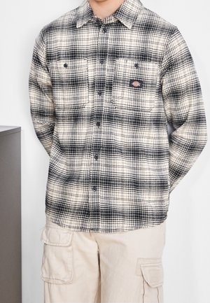 Persona con una camisa de cuadros en blanco y negro abotonada, con dos bolsillos en el pecho, y pantalones cargo beige, de pie frente a un fondo blanco.