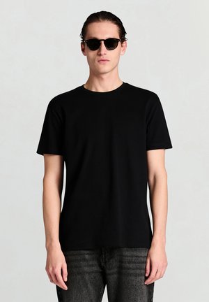 T-shirt nera in cotone con scollo rotondo, maniche corte e vestibilità comoda, abbinata a jeans scuri in denim e occhiali da sole neri.