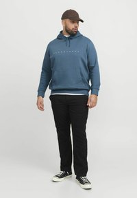 Blauwe hoodie met een voorkruiszak, trekkoord en logo. Gecombineerd met zwarte broek en zwarte sneakers. Model staat tegen een effen achtergrond.