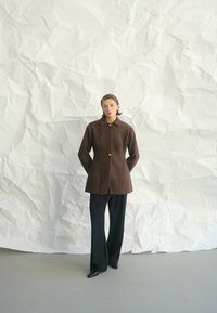 Nő áll egy texturált fehér fal előtt, barna, gombos kabátot, széles szárú fekete nadrágot és fekete hegyes cipőt viselve.